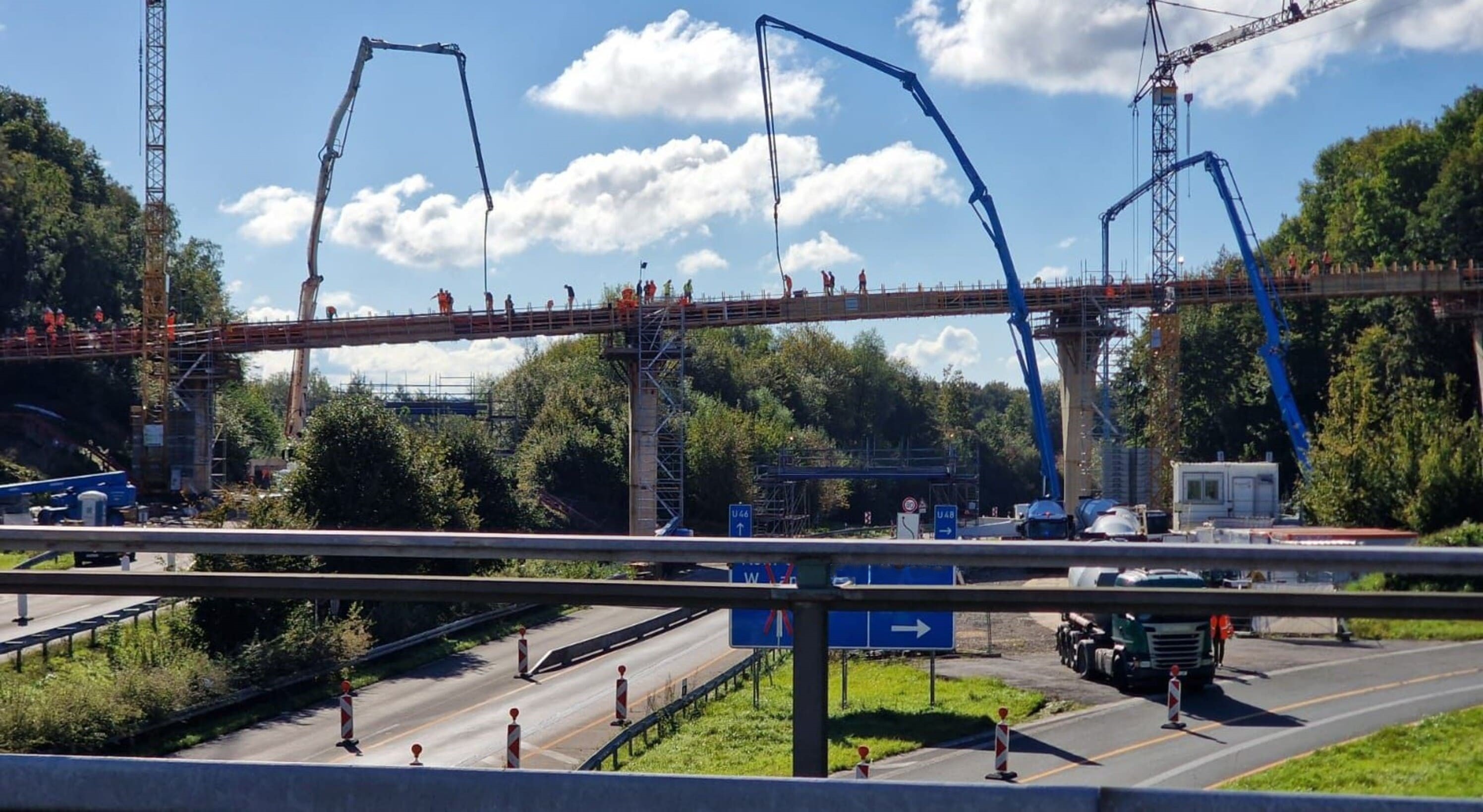Eine im Bau befindliche Brücke überspannt eine mehrspurige Straße. Zahlreiche Bauarbeiter in orangefarbener Schutzkleidung arbeiten auf der Brücke, während mehrere große Betonpumpen mit blauen Auslegern frischen Beton verteilen. Im Vordergrund sind Verkehrszeichen, Baustellenabsperrungen und ein LKW zu sehen. Der Hintergrund ist von Bäumen und einem leicht bewölkten Himmel geprägt.

                                      